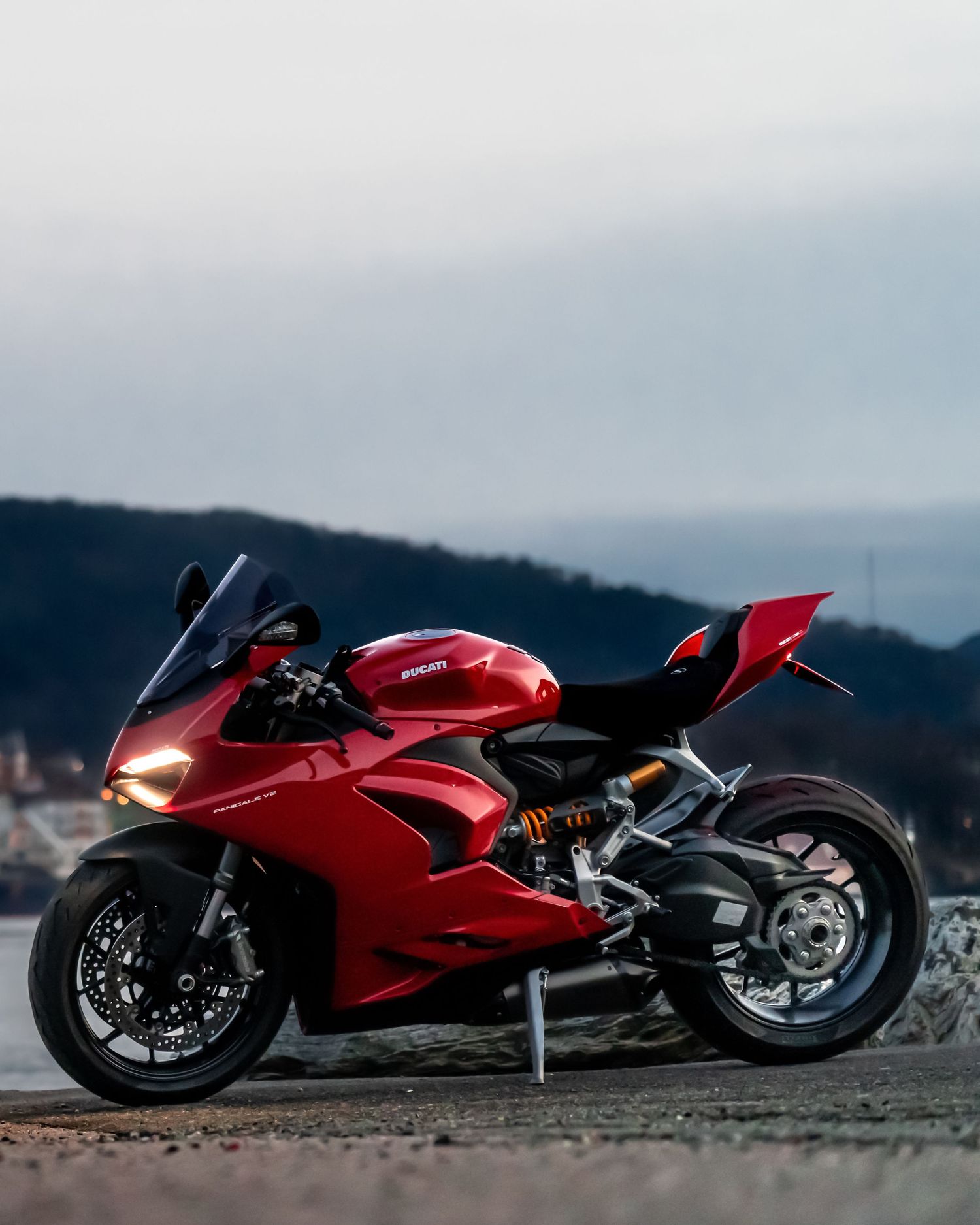 A sleek red Ducati superbike parked against a scenic mountain landscape with overcast skies in the background.