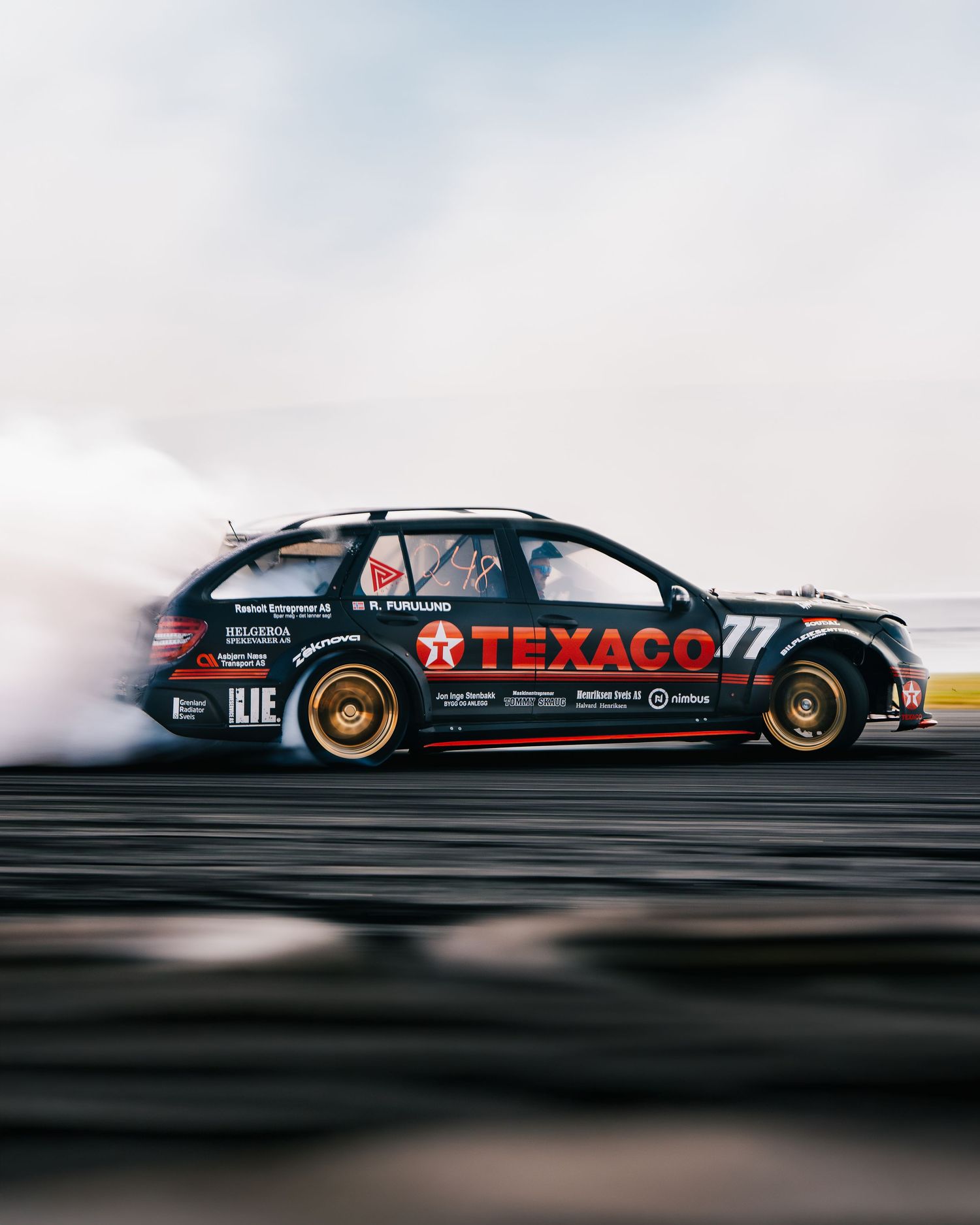 A black racing car with Texaco branding drifts sideways on a dark surface creating a dramatic motion blur effect.
