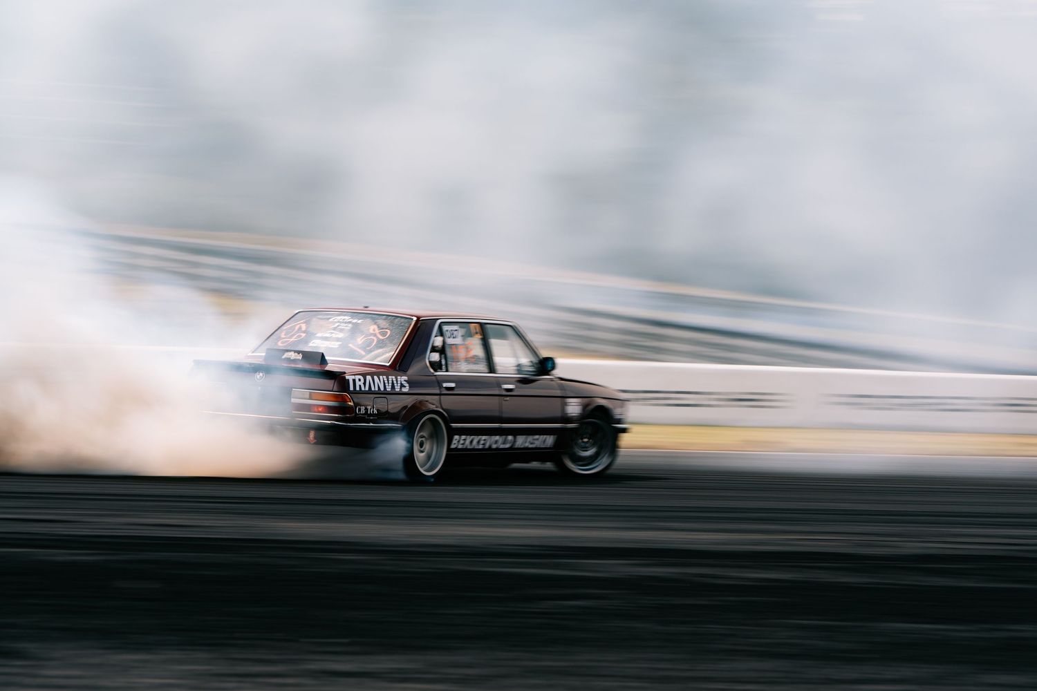 A black car drifts sideways on a racetrack creating a cloud of tire smoke during a motorsport event.
