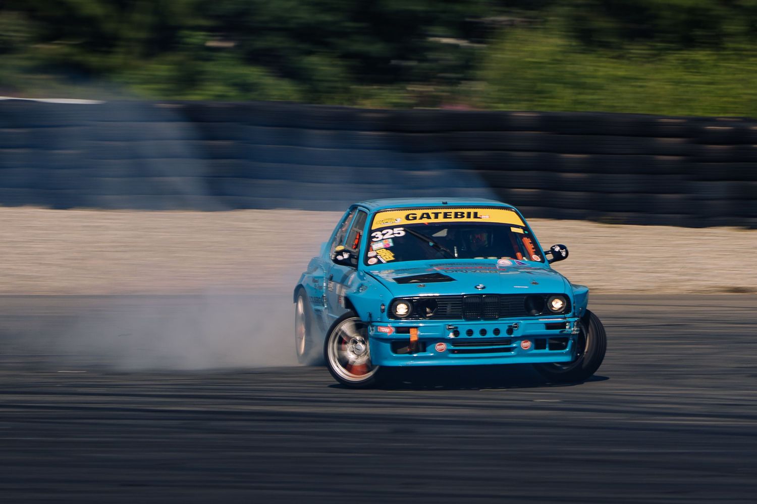 Blue BMW drift car performs a high-speed slide during a drift competition, creating smoke from spinning tires.