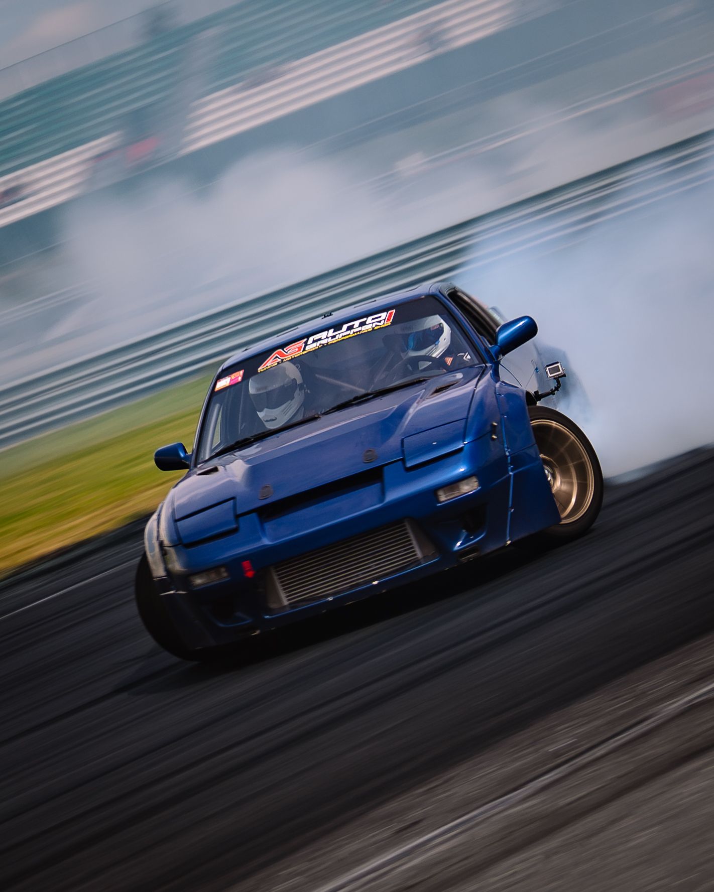 A blue sports car drifts sideways on a racetrack creating dramatic smoke trails during a drift competition.