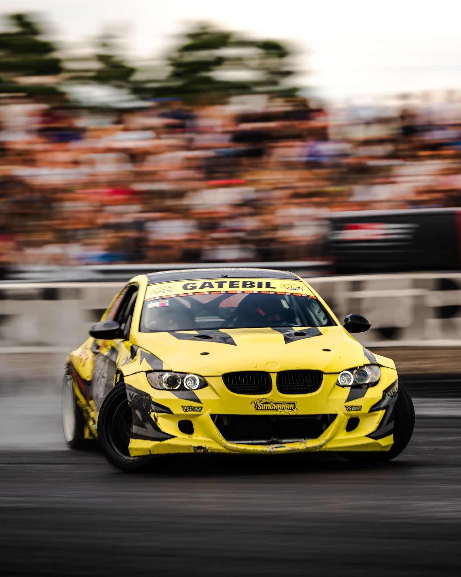 Yellow BMW drift cars sliding in formation across racetrack with motion blur capturing dynamic action.