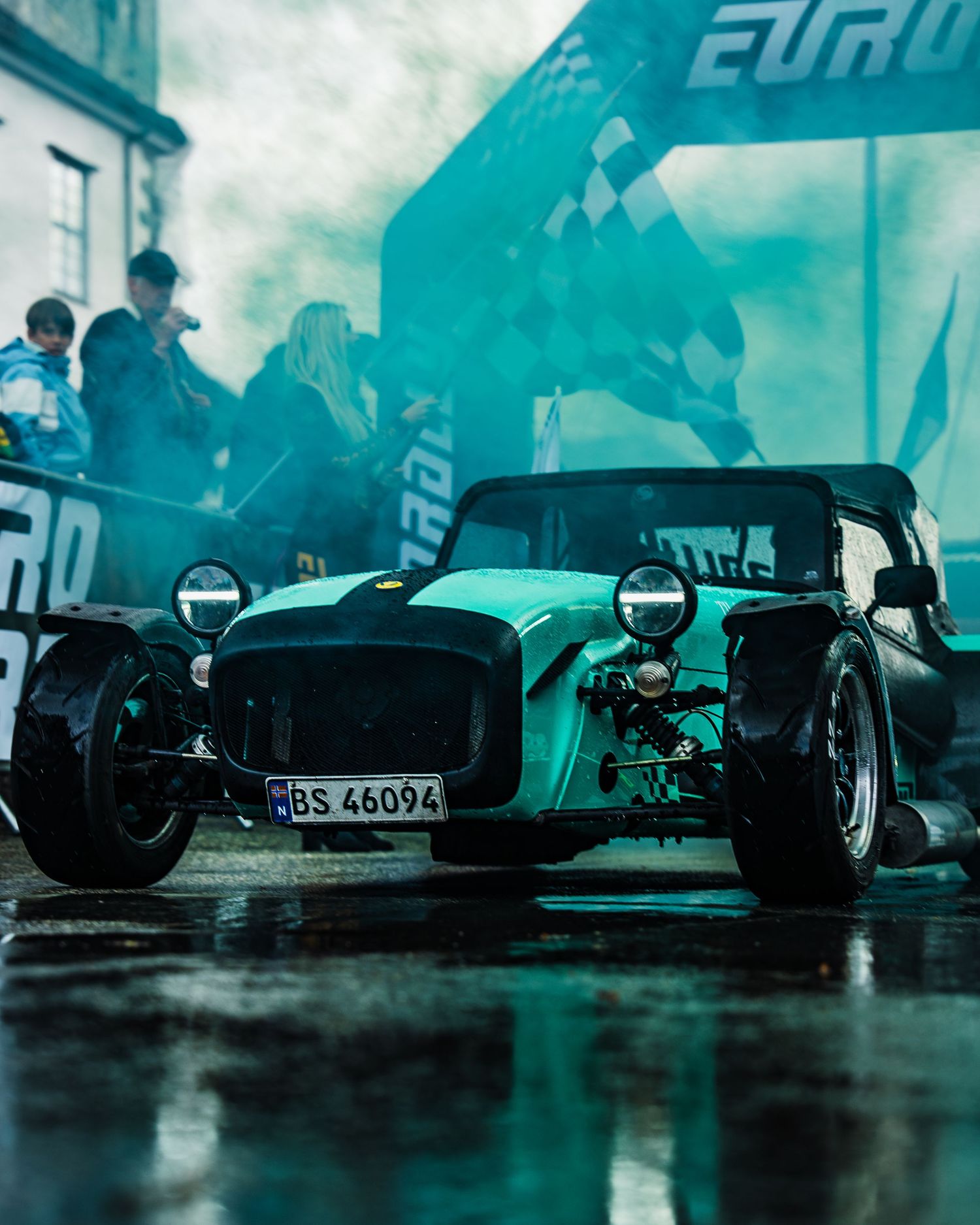 Vintage Caterham race car under turquoise lighting at EuroRally event in wet conditions.
