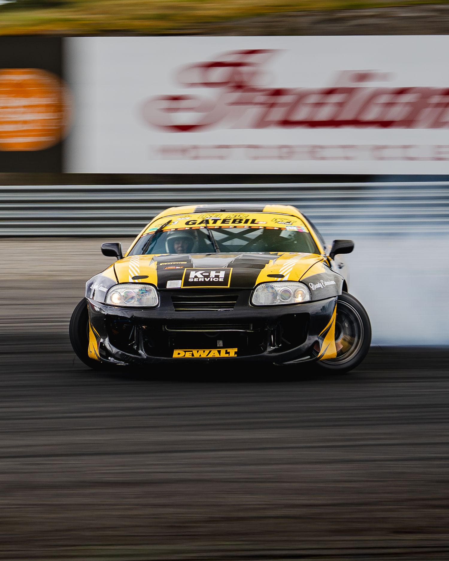 Black and yellow modified Toyota Supra drifting on a race track with motion blur and smoke from the tires.