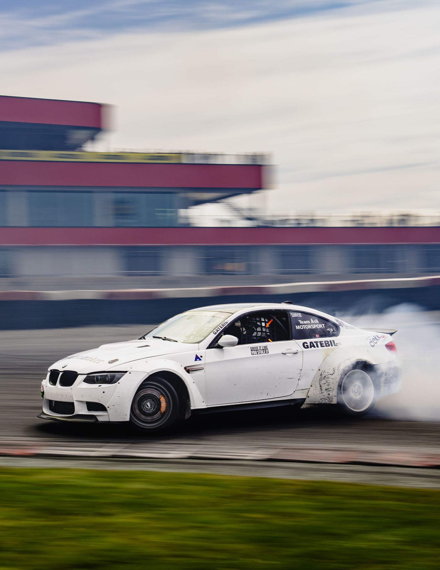 A white BMW race car drifts sideways on a track creating dramatic smoke trails during a professional drifting event.