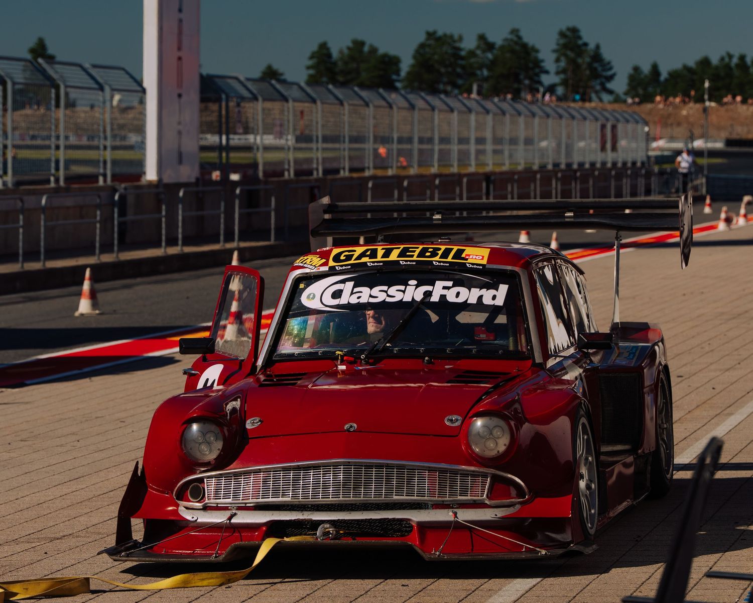 A vintage red Classic Ford race car sits parked in pit lane at a motorsport track during sunset.