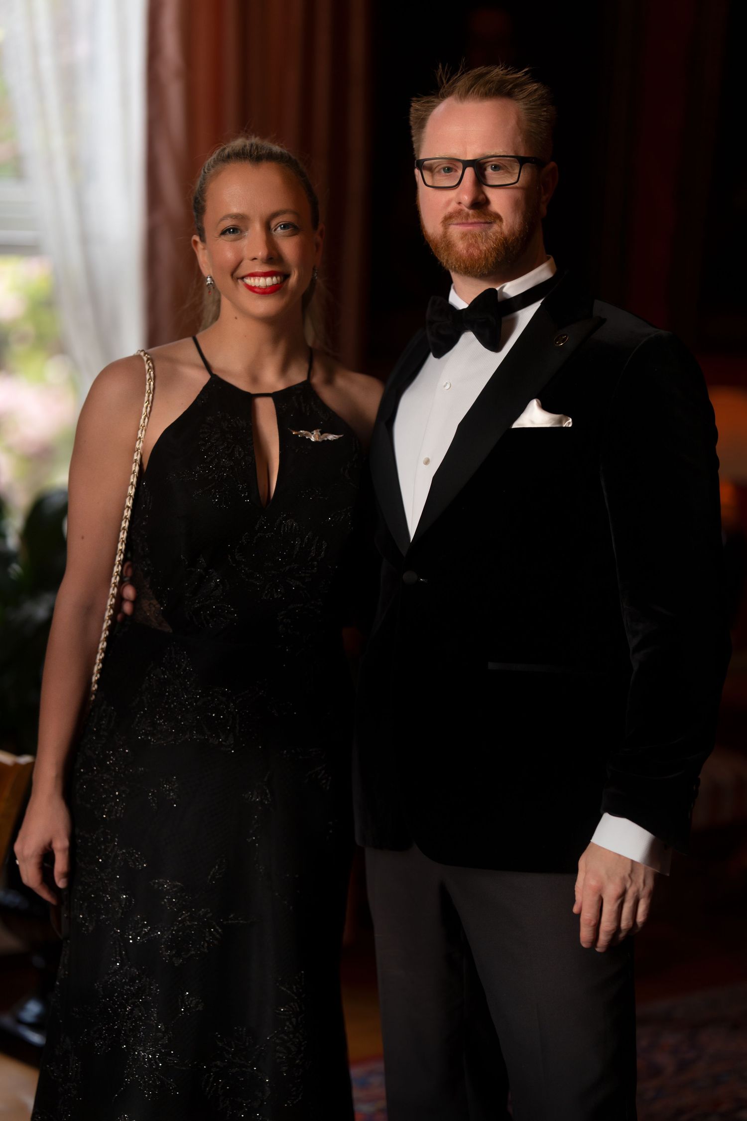 Elegant couple posing at formal event, with her in a sparkly black gown and him in a black tuxedo.