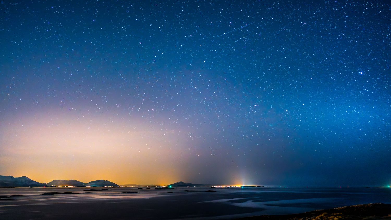Night sky panorama showing stars and city lights along coastal horizon.