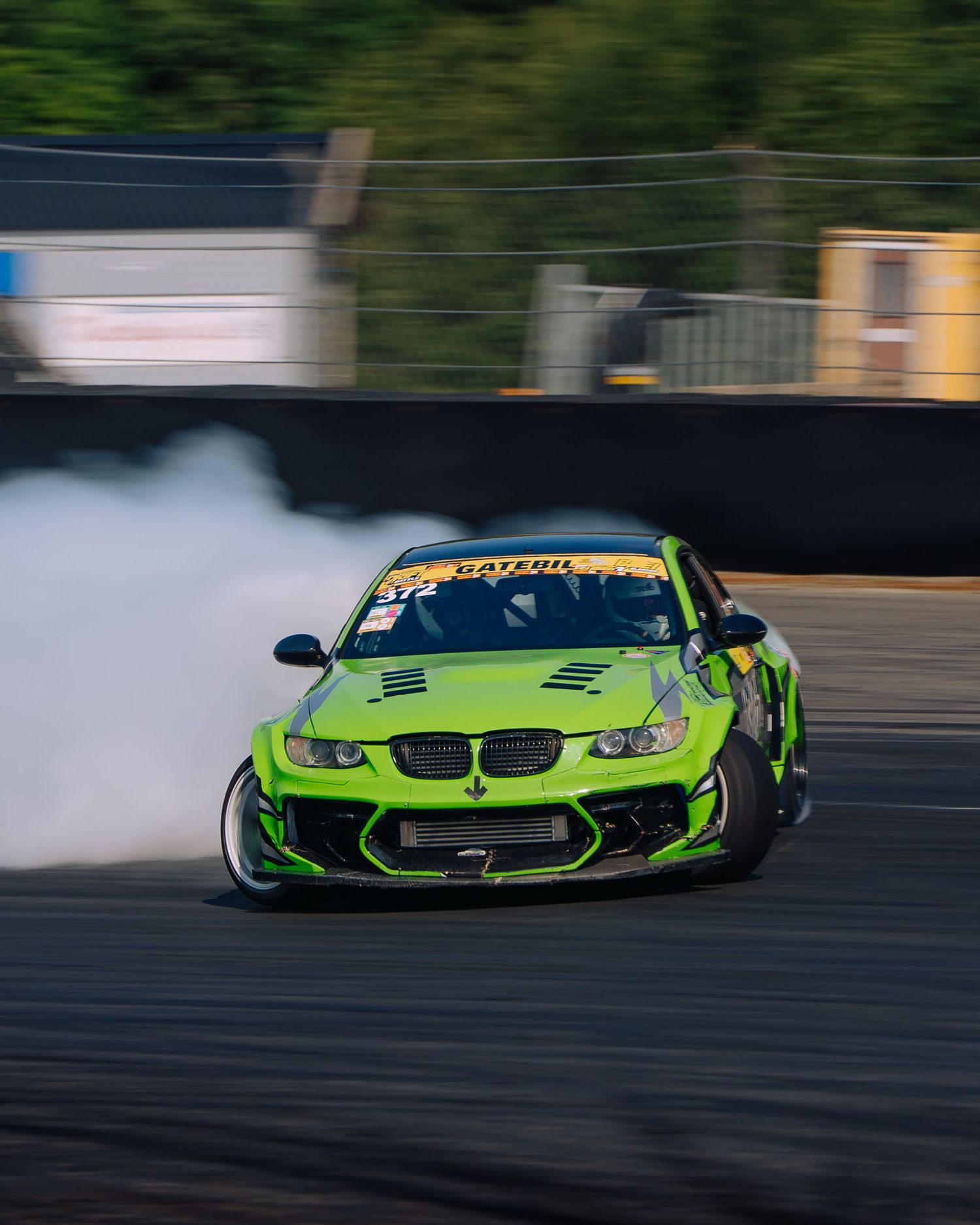 A green BMW race car drifts sideways on a track with tire smoke billowing behind it.