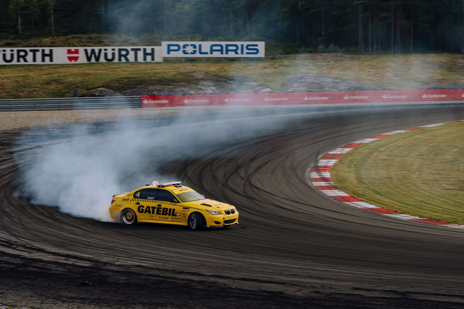 Yellow drift car creates smoke while sliding through a corner on a race track with Wurth and Polaris signage visible.