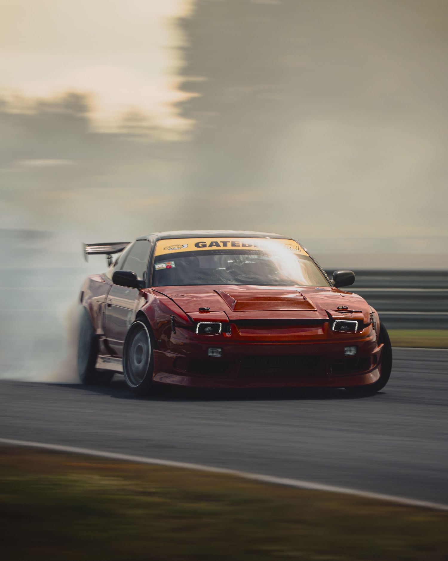 A red sports car drifts sideways on a racetrack creating smoke from its tires during sunset.
