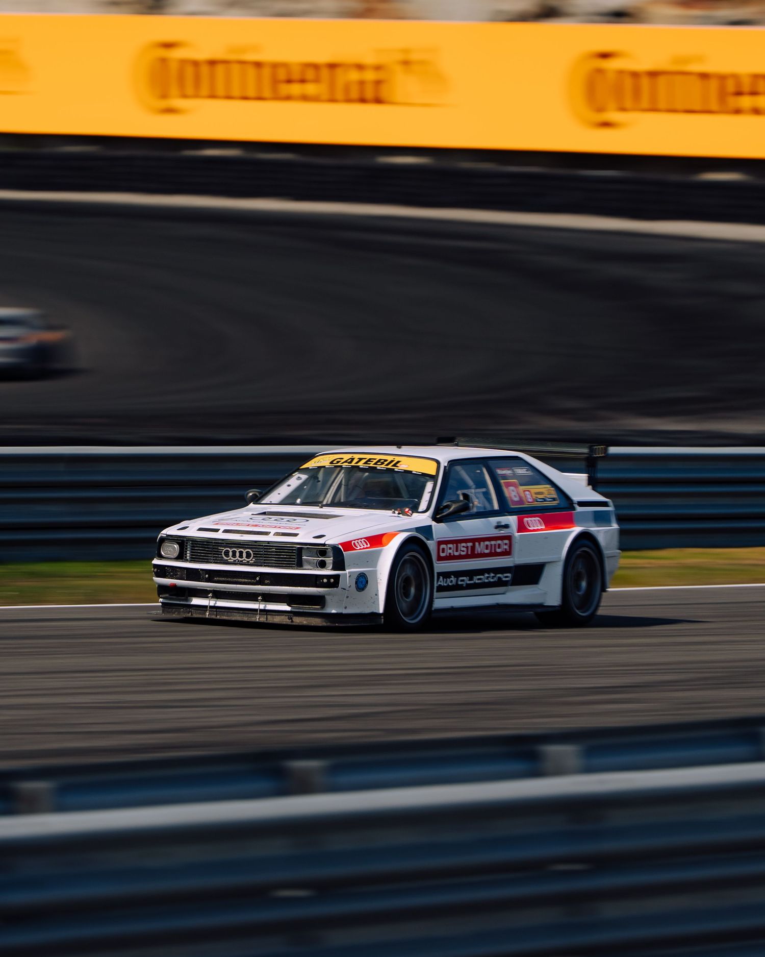 Two vintage race cars speed along a racetrack at sunset with dramatic motion blur and golden lighting.