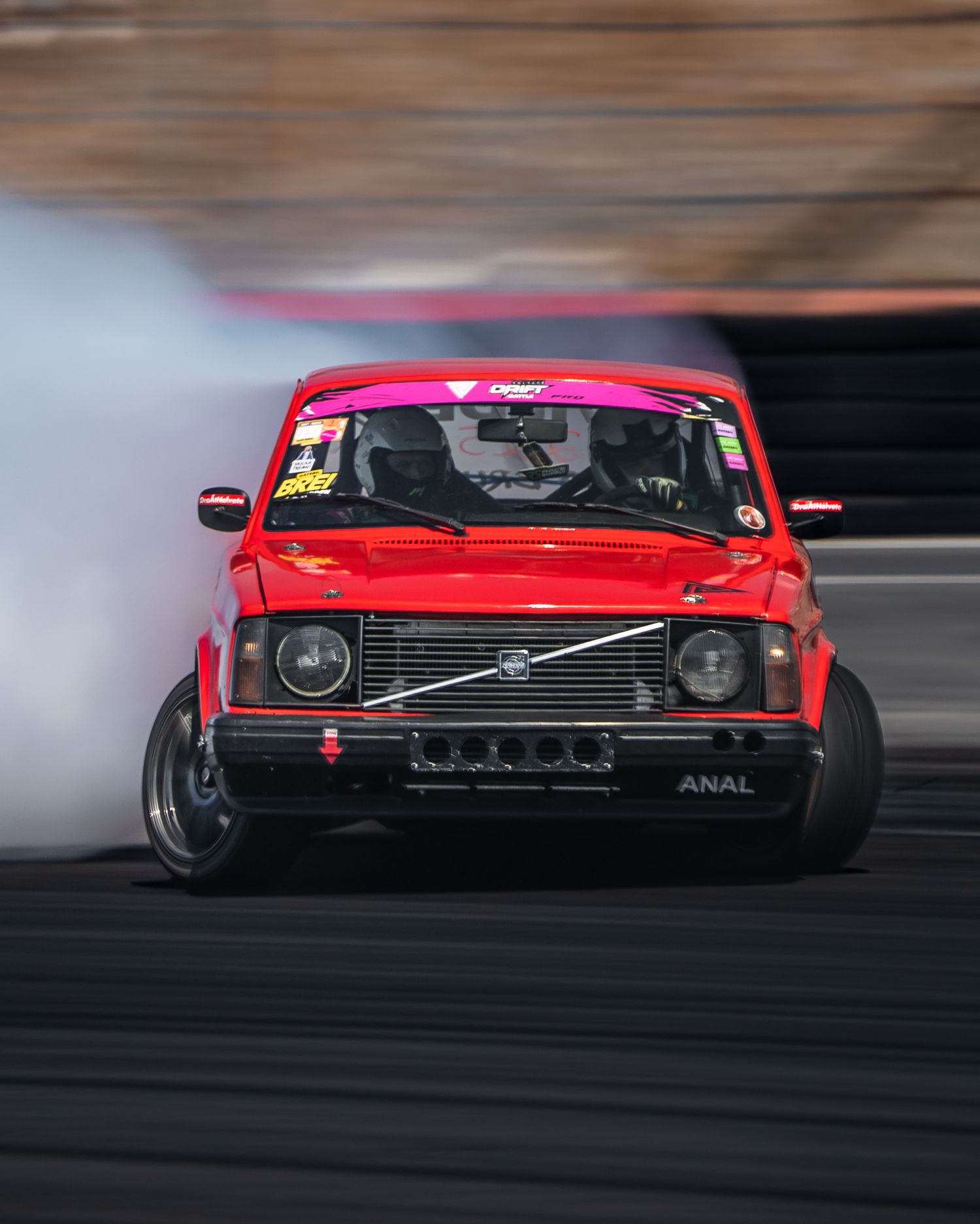 A red drift car slides sideways on a racetrack creating smoke from its tires during a drifting competition.