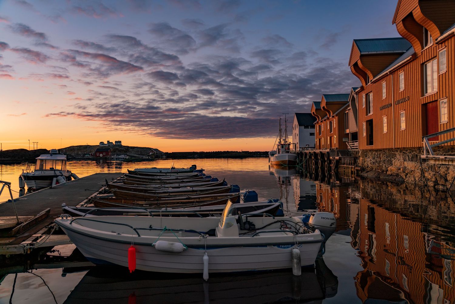 Wooden boats moored along waterfront with orange buildings reflecting in calm harbor at sunset.