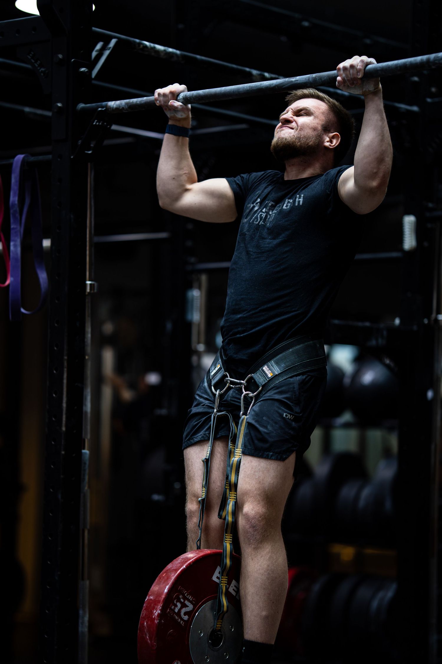 Athletic figure doing strict pull-ups with weight plate attached in dark gym environment.