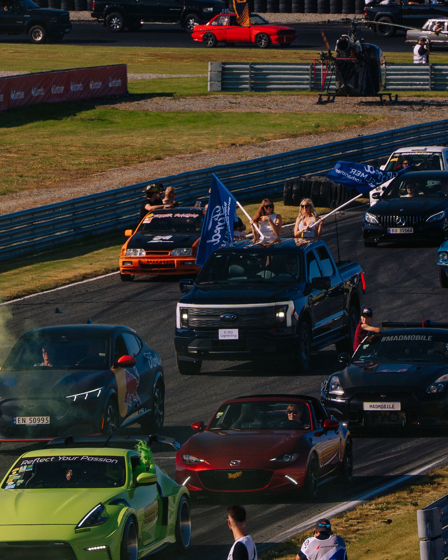 Colorful sports cars lined up at a racetrack event with spectators watching from behind barriers.