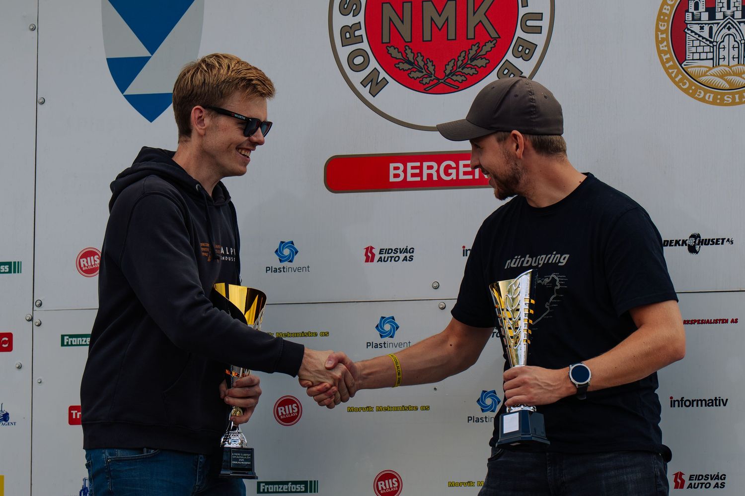 Two people wearing black t-shirts shake hands at a professional event with sponsor logos visible in the background.