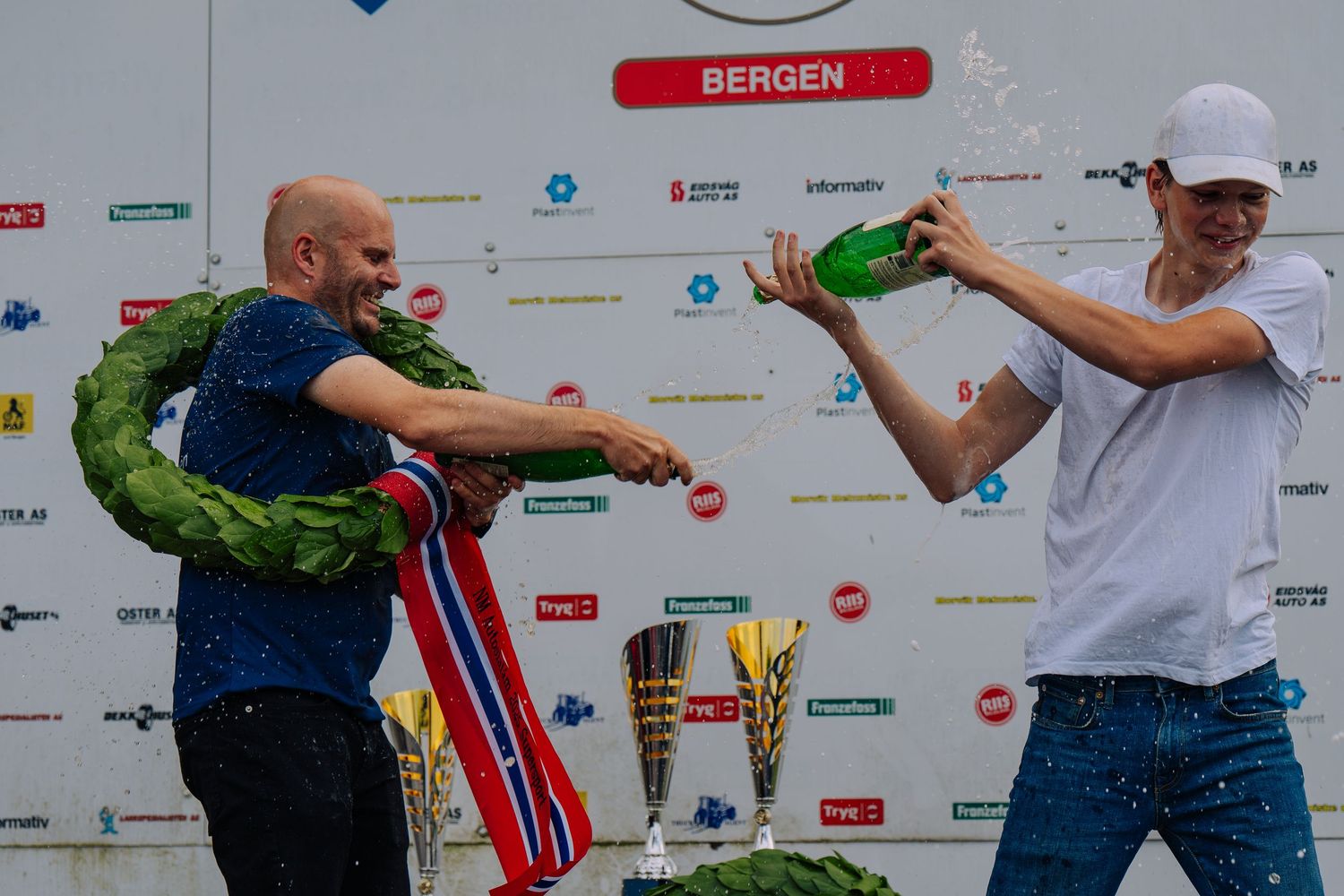 Athletes celebrate at the finish line of a sports competition in Bergen, Norway with sponsor banners visible in background.