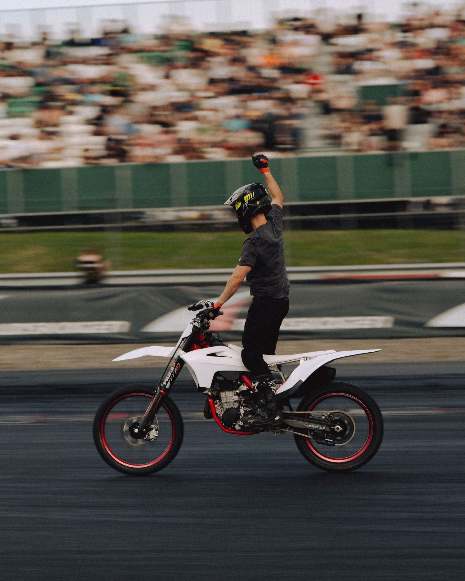 A motorcycle stunt rider performs a trick on a white dirt bike at a racing track with blurred spectators in the background.