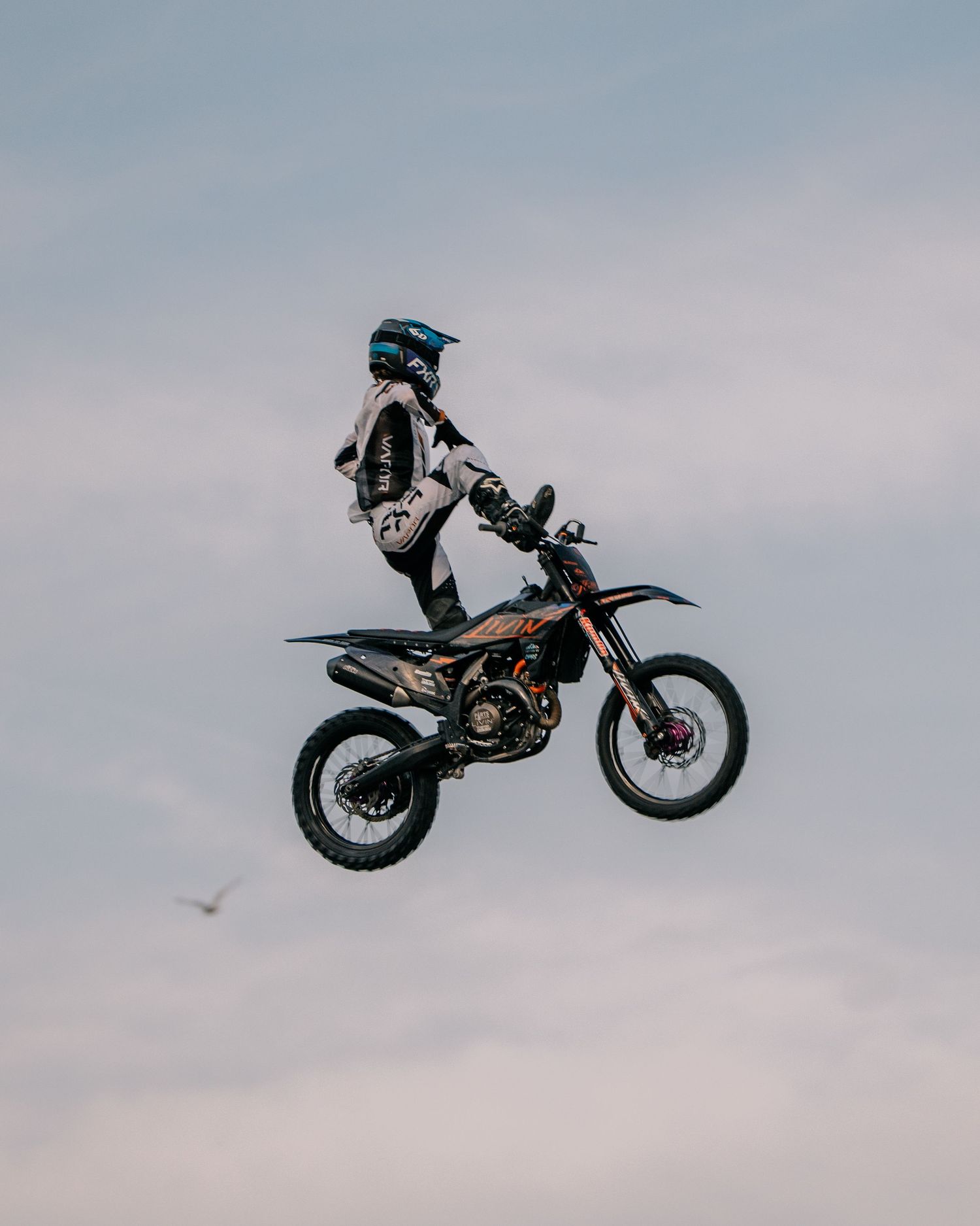 Motocross rider performing a dramatic aerial stunt jump against an overcast sky.