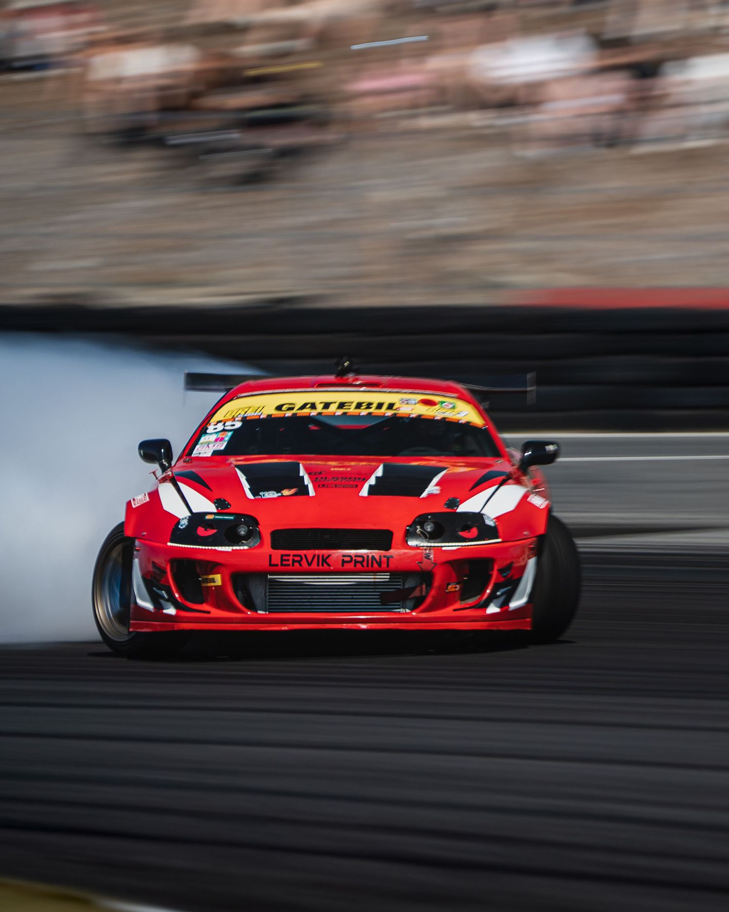 Red race car drifts on track creating smoke while cornering at high speed during motorsport competition.