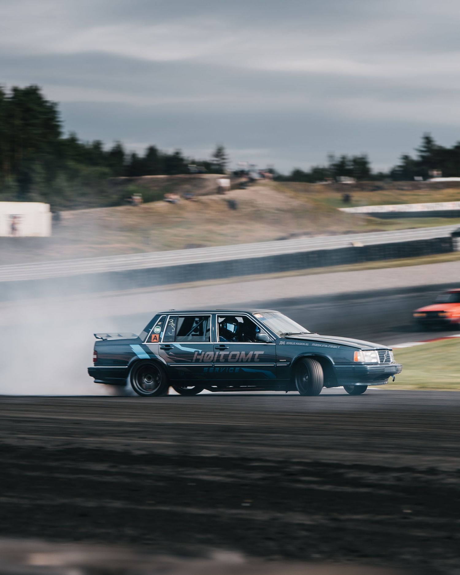 A dark blue Lincoln Town Car drifts sideways on a race track with smoke trailing from its tires.