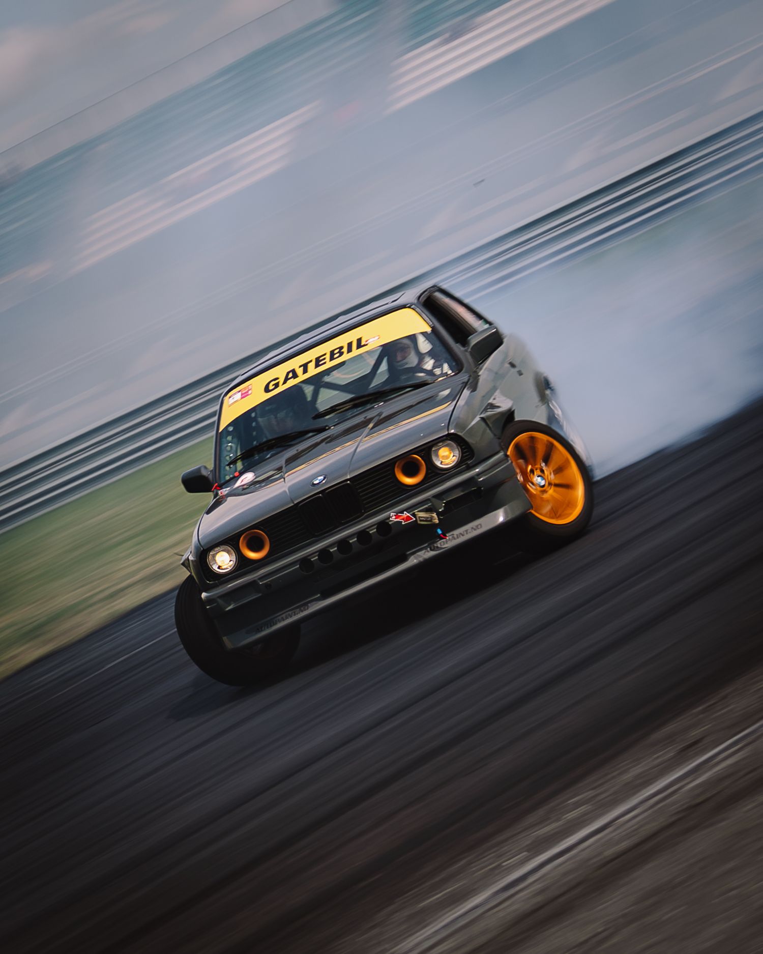 Black sports car drifting on racetrack with orange wheels creating smoke during motorsport competition.