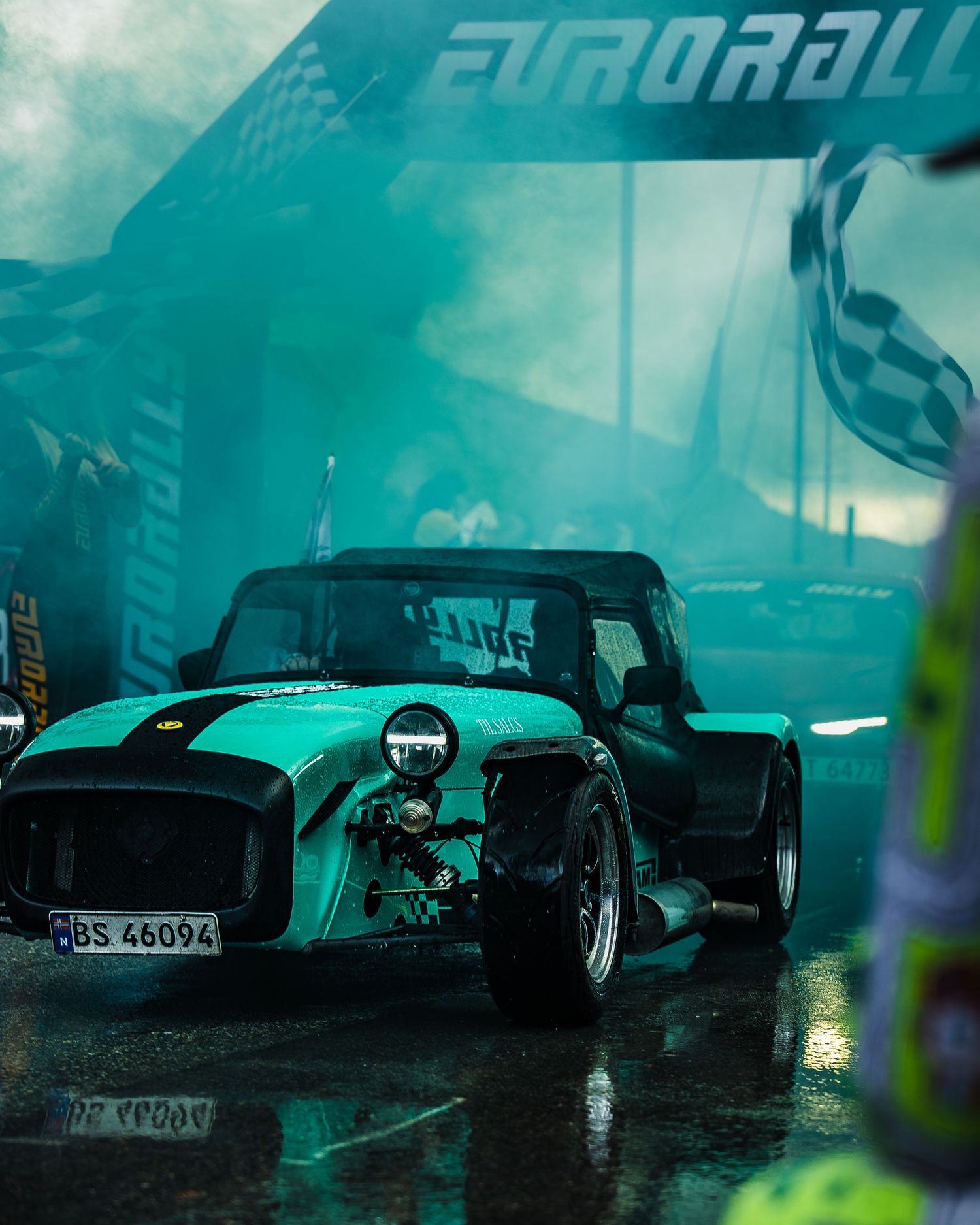 Classic Caterham sports car illuminated by teal lighting at rainy motorsport event.