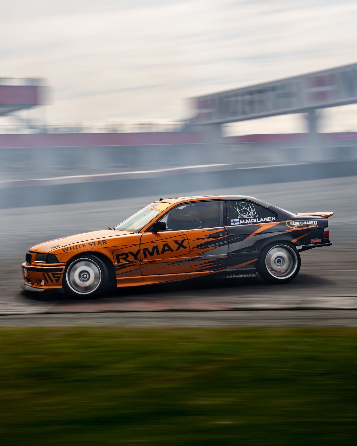 Orange BMW drift car performing a smoky drift maneuver on a racetrack with motion blur effect.