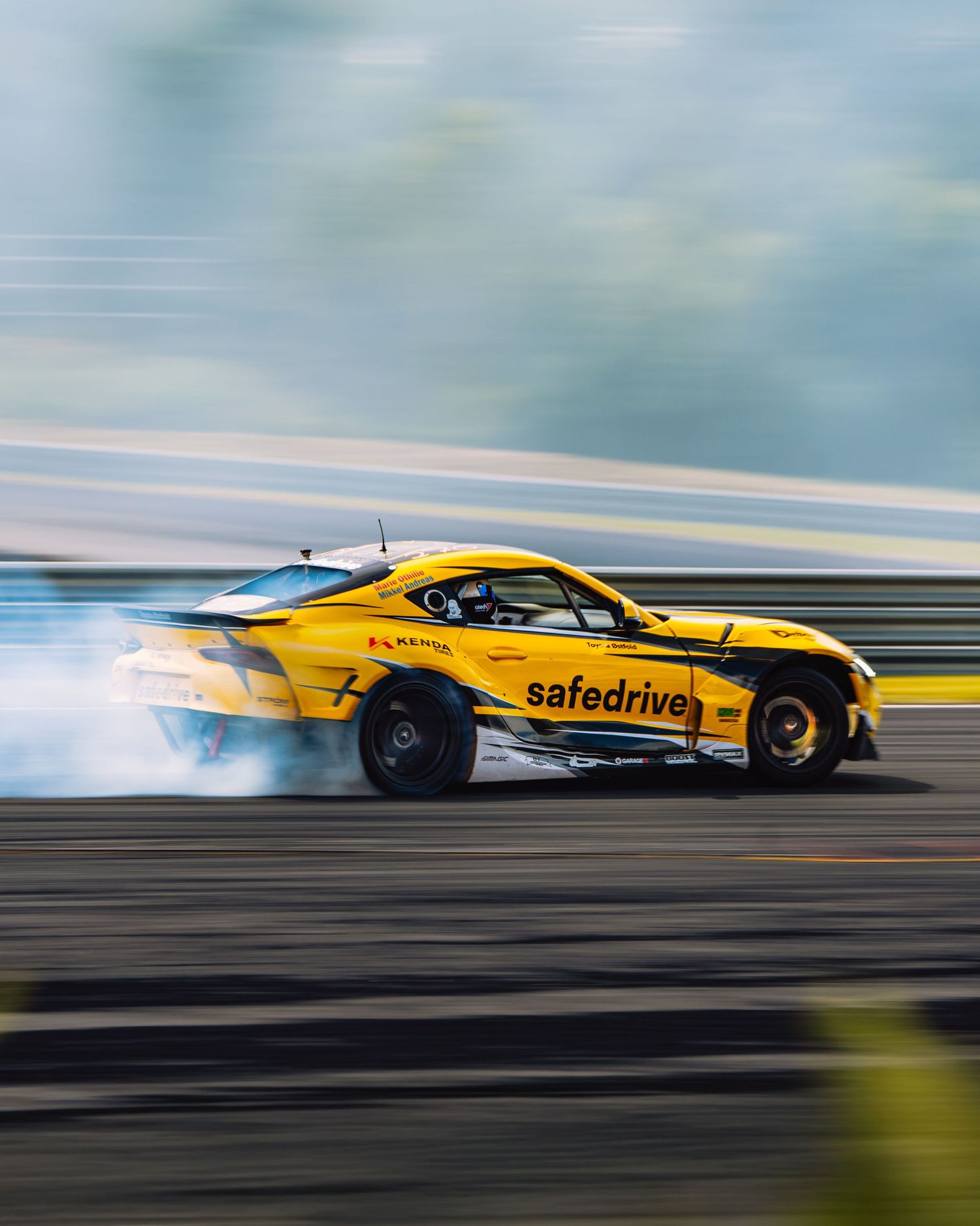 A yellow drift car slides sideways on a race track creating smoke while performing a controlled drift maneuver.