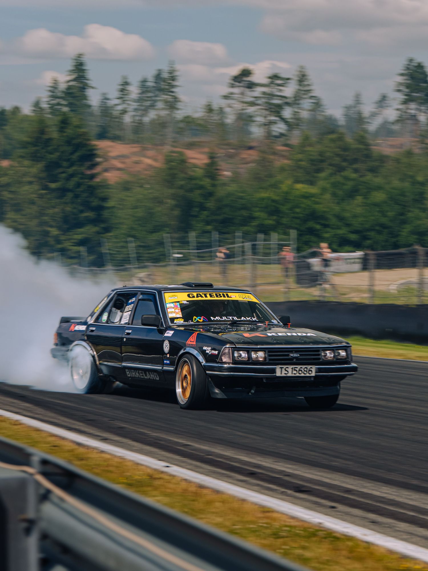 Black drift car performs a smoky tandem drift run on a racetrack with evergreen trees in the background.