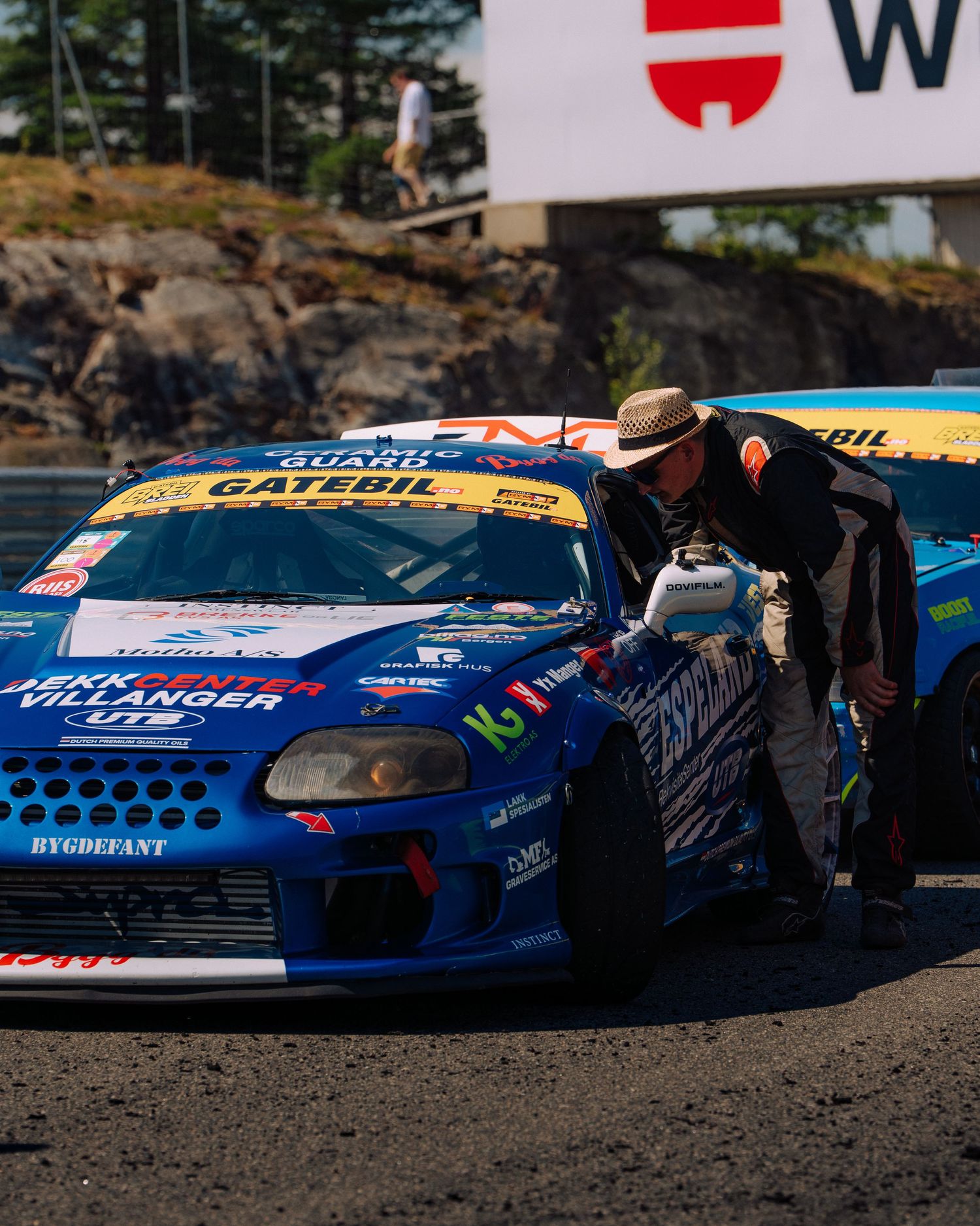A blue racing car with Gate Oil sponsorship being inspected by a person in a hat at a racetrack.