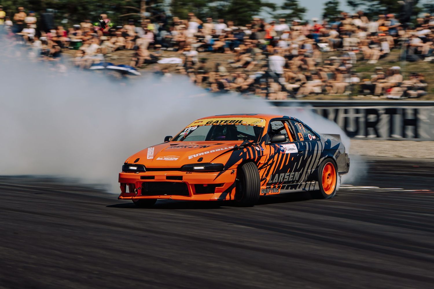 Orange drift car slides sideways on racetrack creating smoke during competitive drifting event.