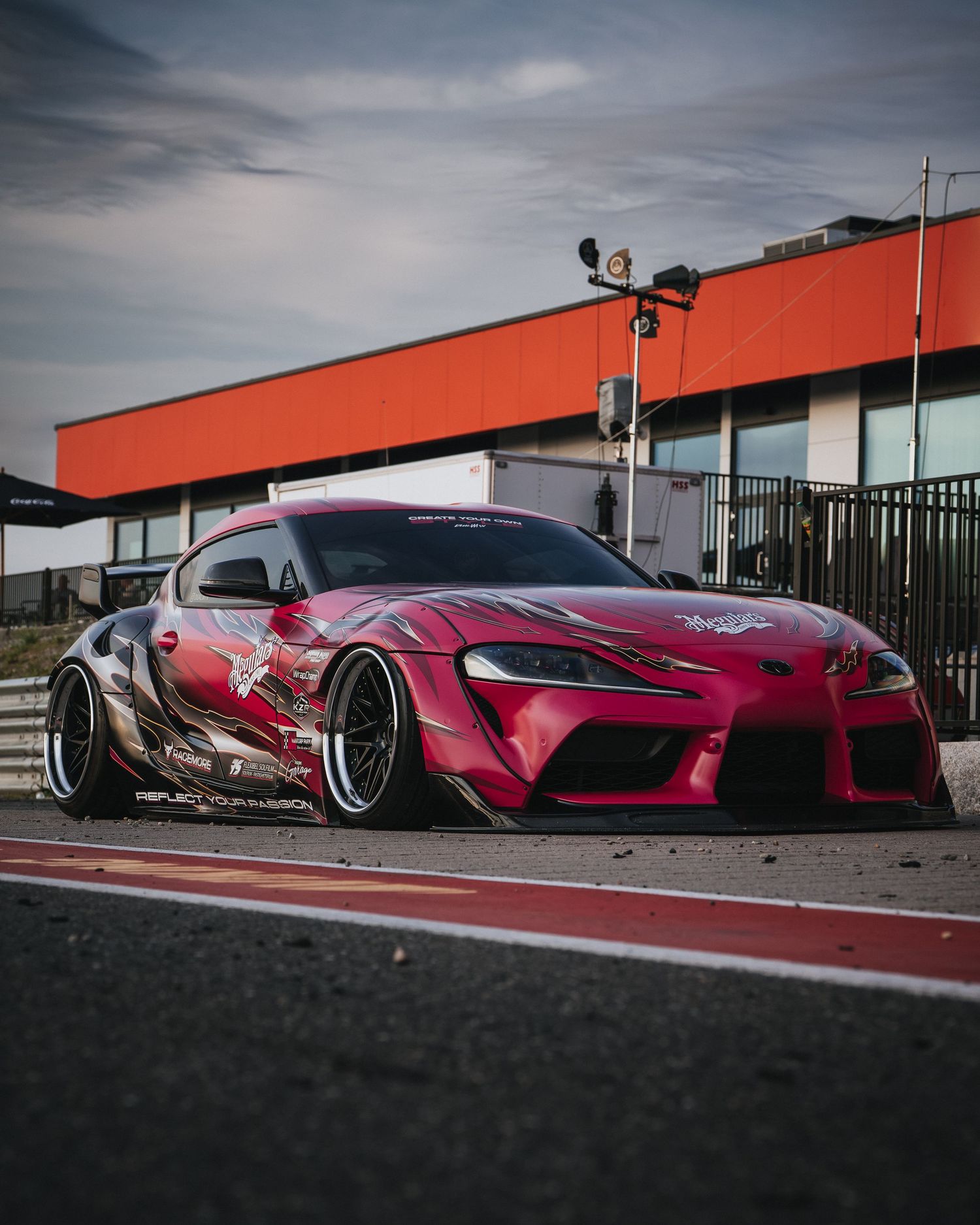 A modified red Toyota Supra with wide body kit and custom graphics sits low on a race track against an industrial backdrop.