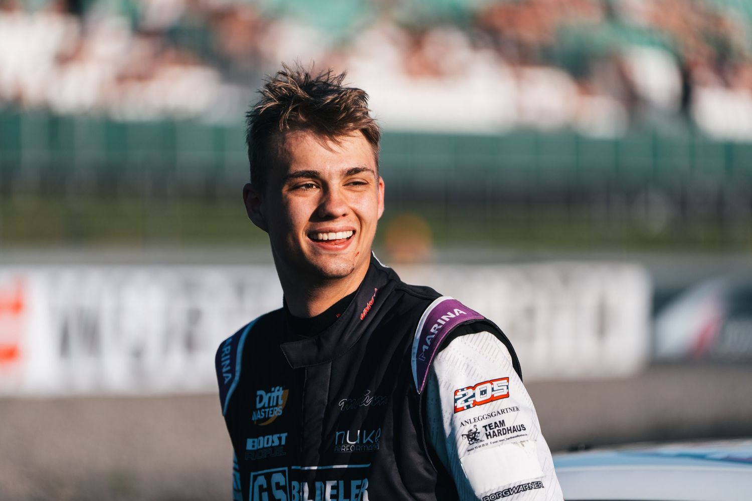 Racing driver in purple and white racing suit at a motorsport event.