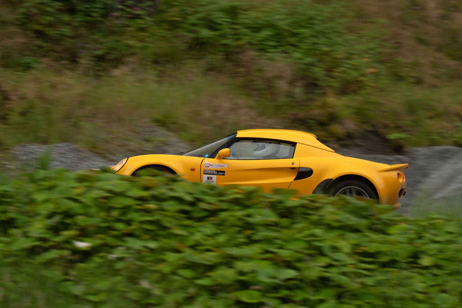 A yellow Lotus sports car drives through a winding mountain road surrounded by lush green foliage.