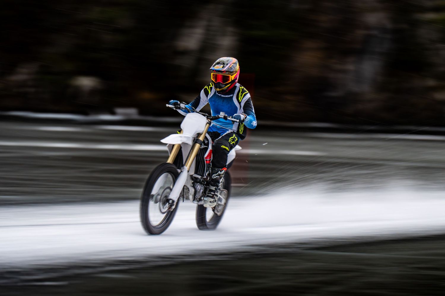 A motorcycle rider performs a dramatic wheelie stunt on a snowy winter road, creating a dynamic motion blur effect.