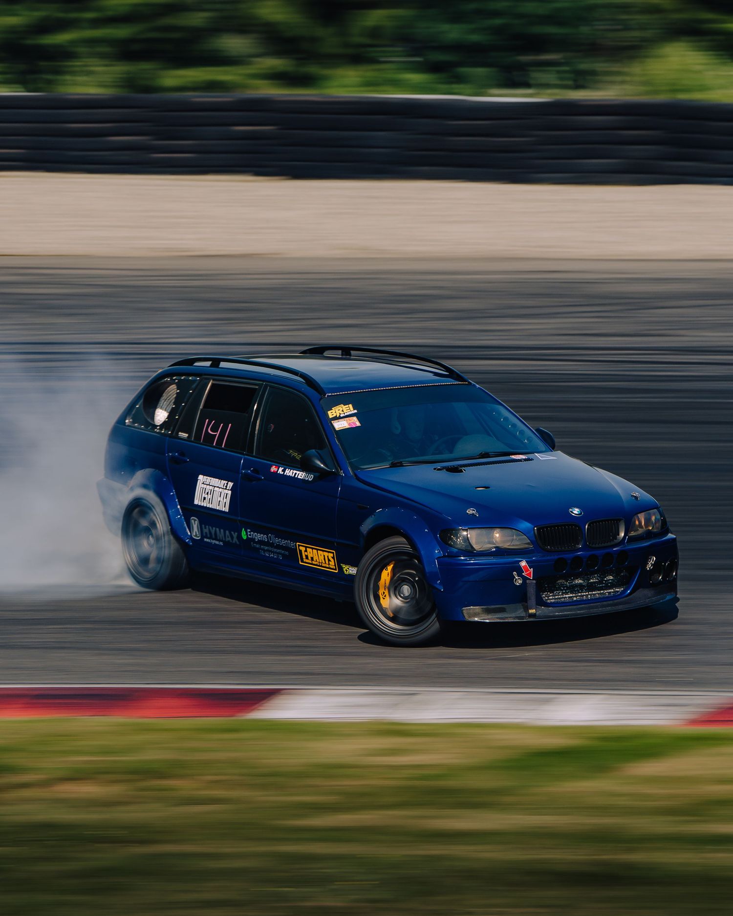 Blue modified BMW drifting on a racetrack with motion blur and smoke from the tires.