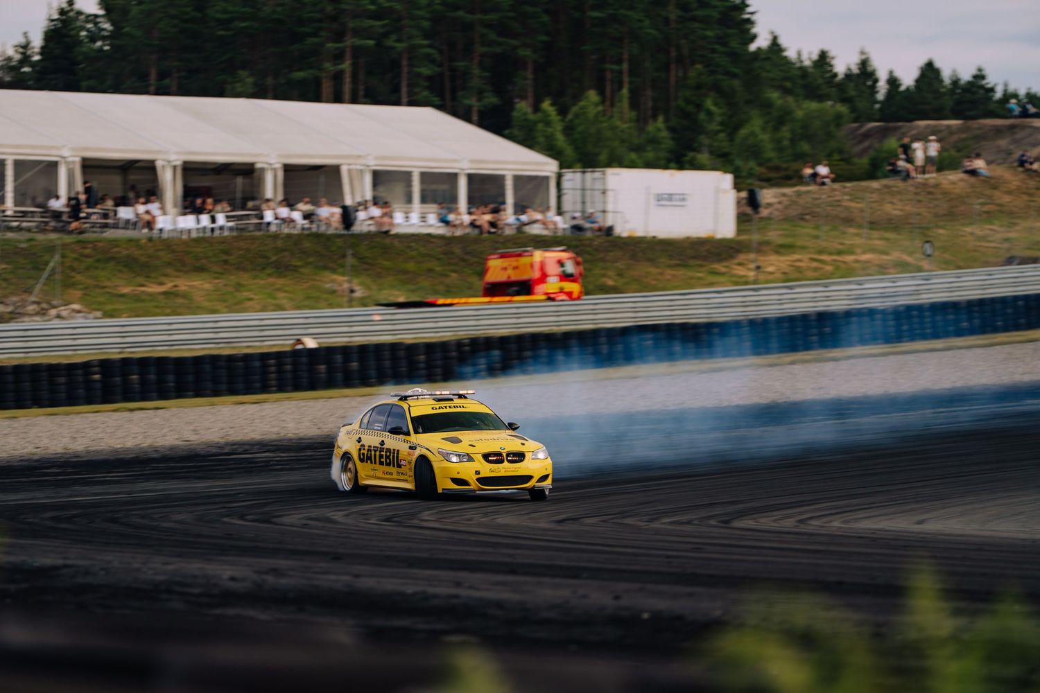 Yellow drift car creates smoke while drifting on a racetrack with evergreen trees in the background.