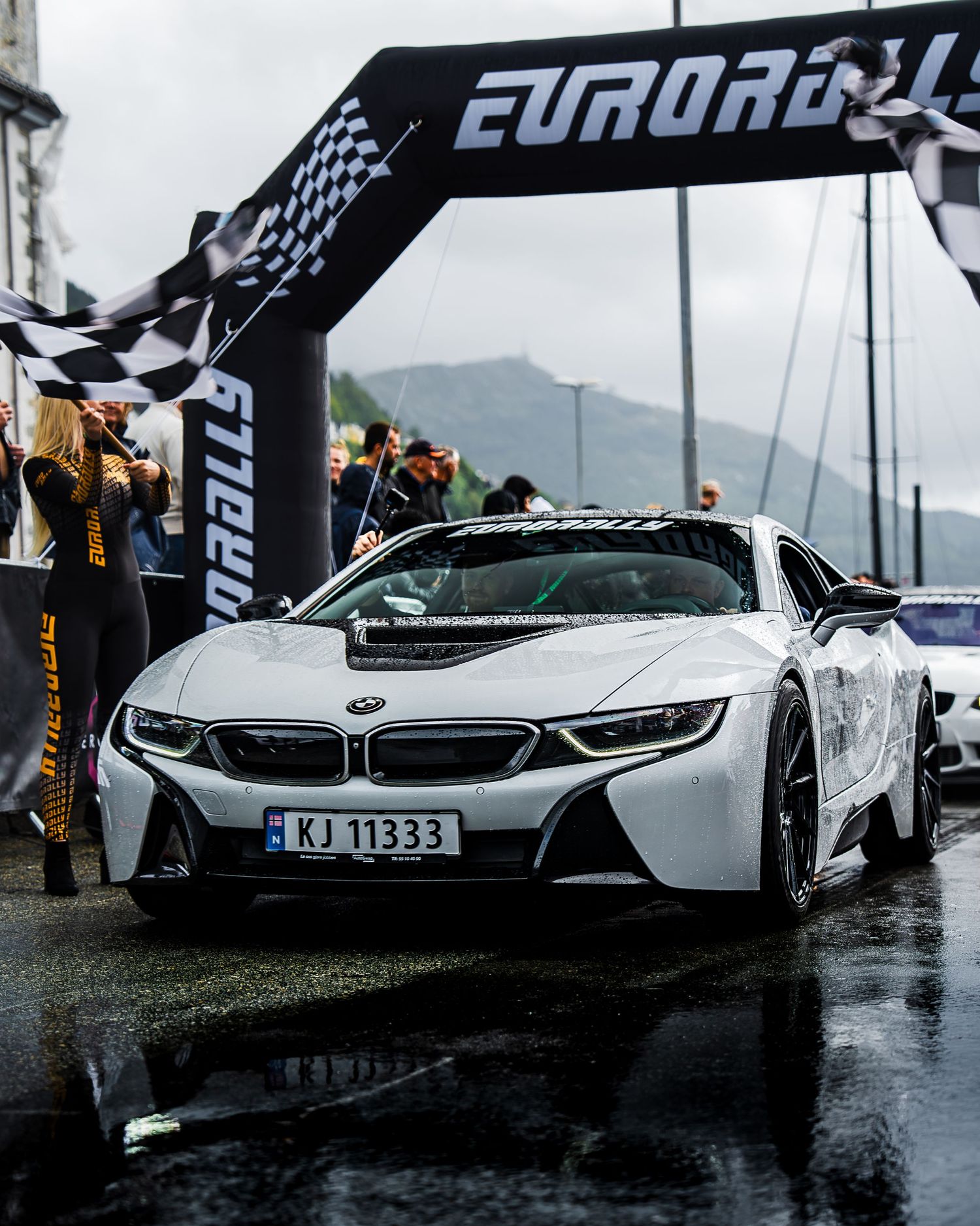 White BMW i8 hybrid supercar crossing under Eurorally arch during rainy race event.