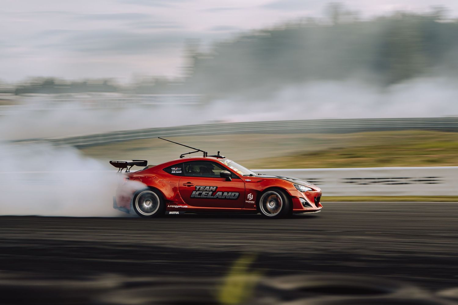 Red drift car creates smoke clouds while performing on race track during motorsport competition.