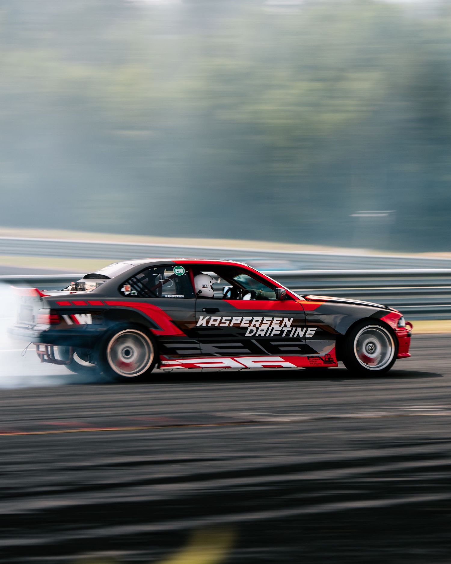 Red and black drift car creating smoke while drifting sideways on a racetrack during high-speed motorsport action.