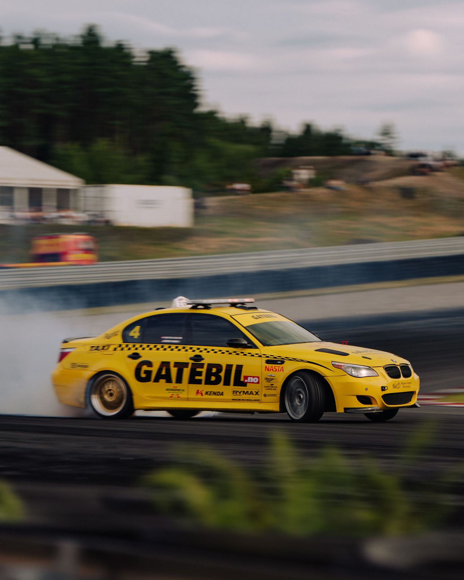 Yellow BMW drift car speeds past on a racetrack with motion blur effect.