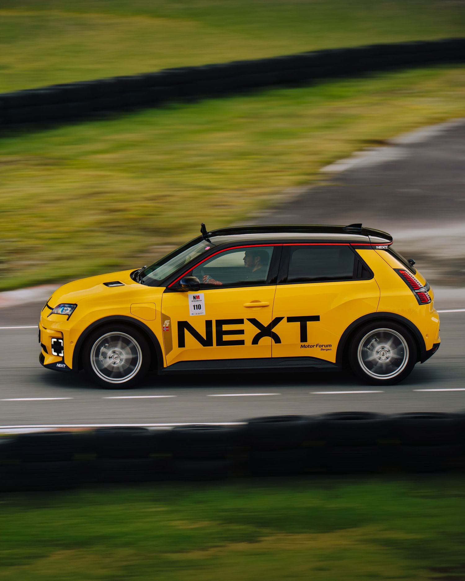 A bright yellow electric car with 'NEXT' branding in motion on a curved road against a green background.
