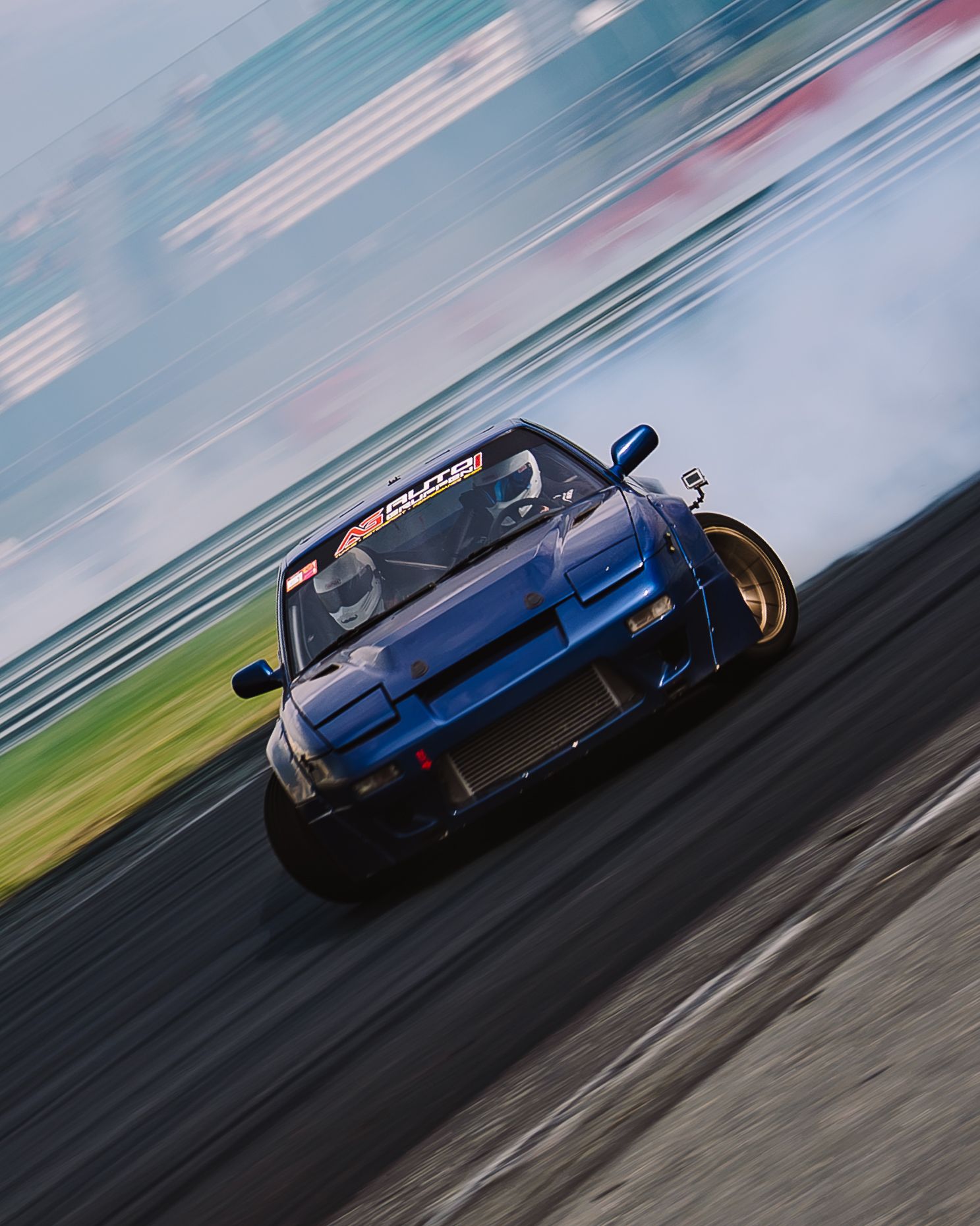 A blue sports car drifts sideways on a racetrack creating smoke from its tires during a drifting competition.