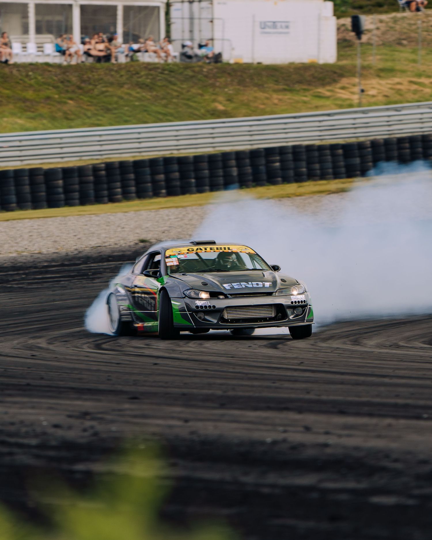 A black drift car slides sideways on a racetrack creating smoke from burning rubber during a drift competition.