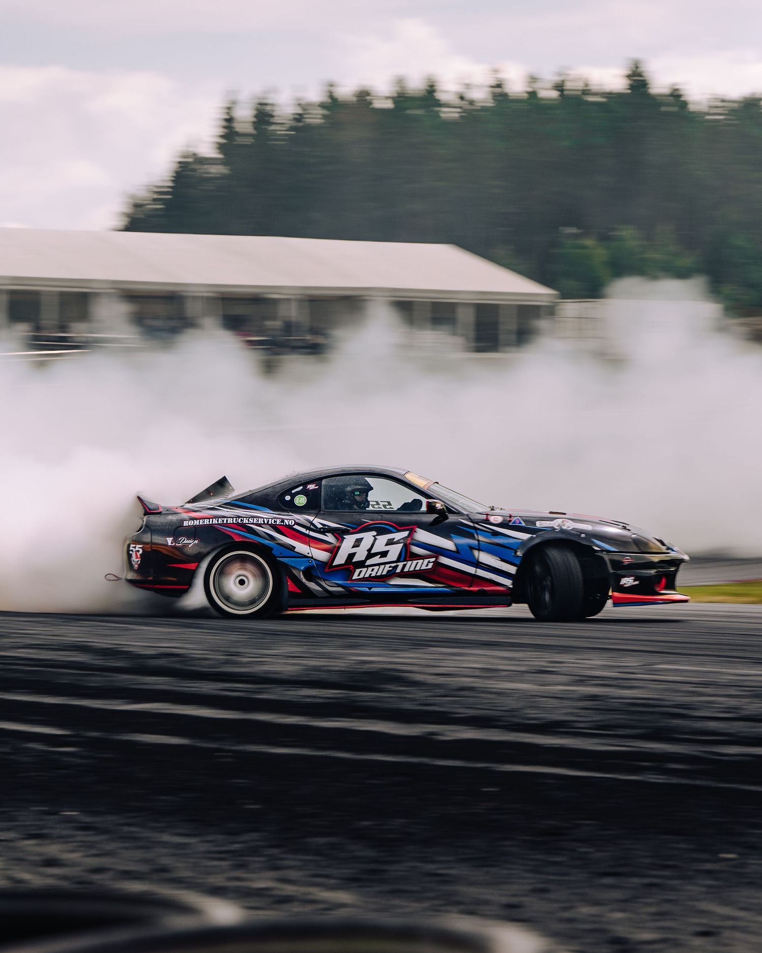 A racing car drifts sideways on the track creating a cloud of tire smoke during a motorsport competition.