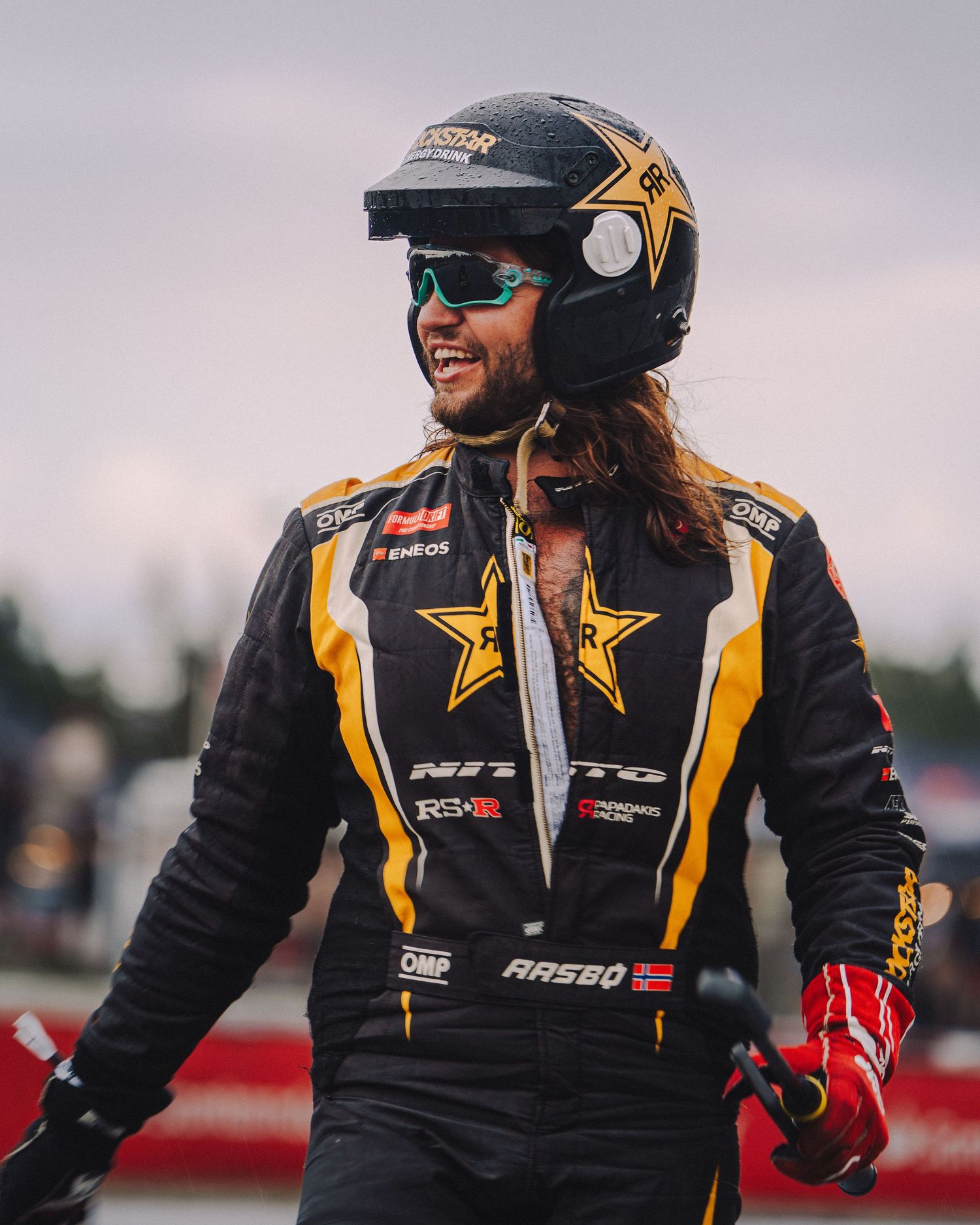 Racing driver in black and yellow uniform jacket and sunglasses celebrates on track with energetic expressions.