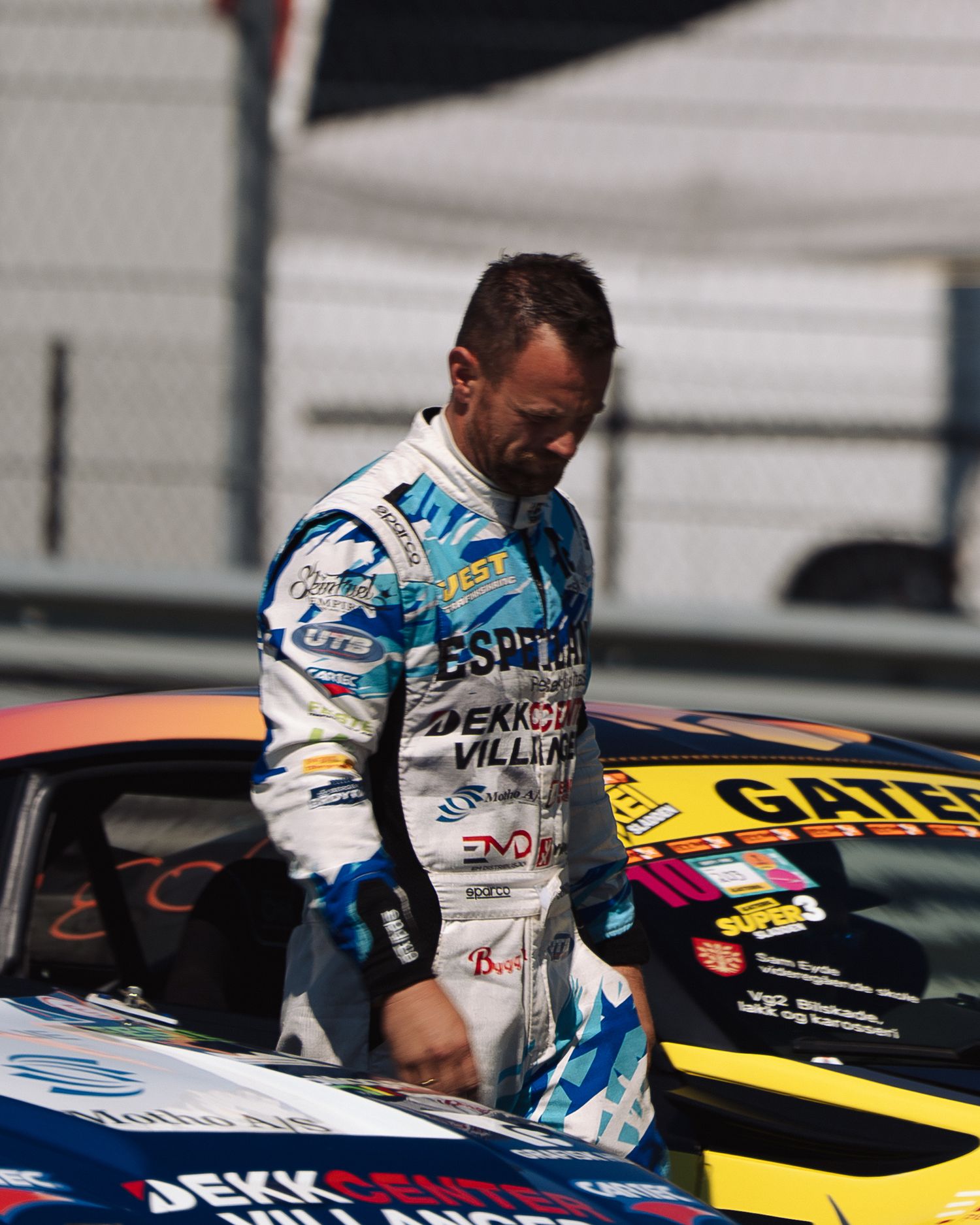NASCAR driver in white racing suit climbs into yellow race car through window at speedway track.