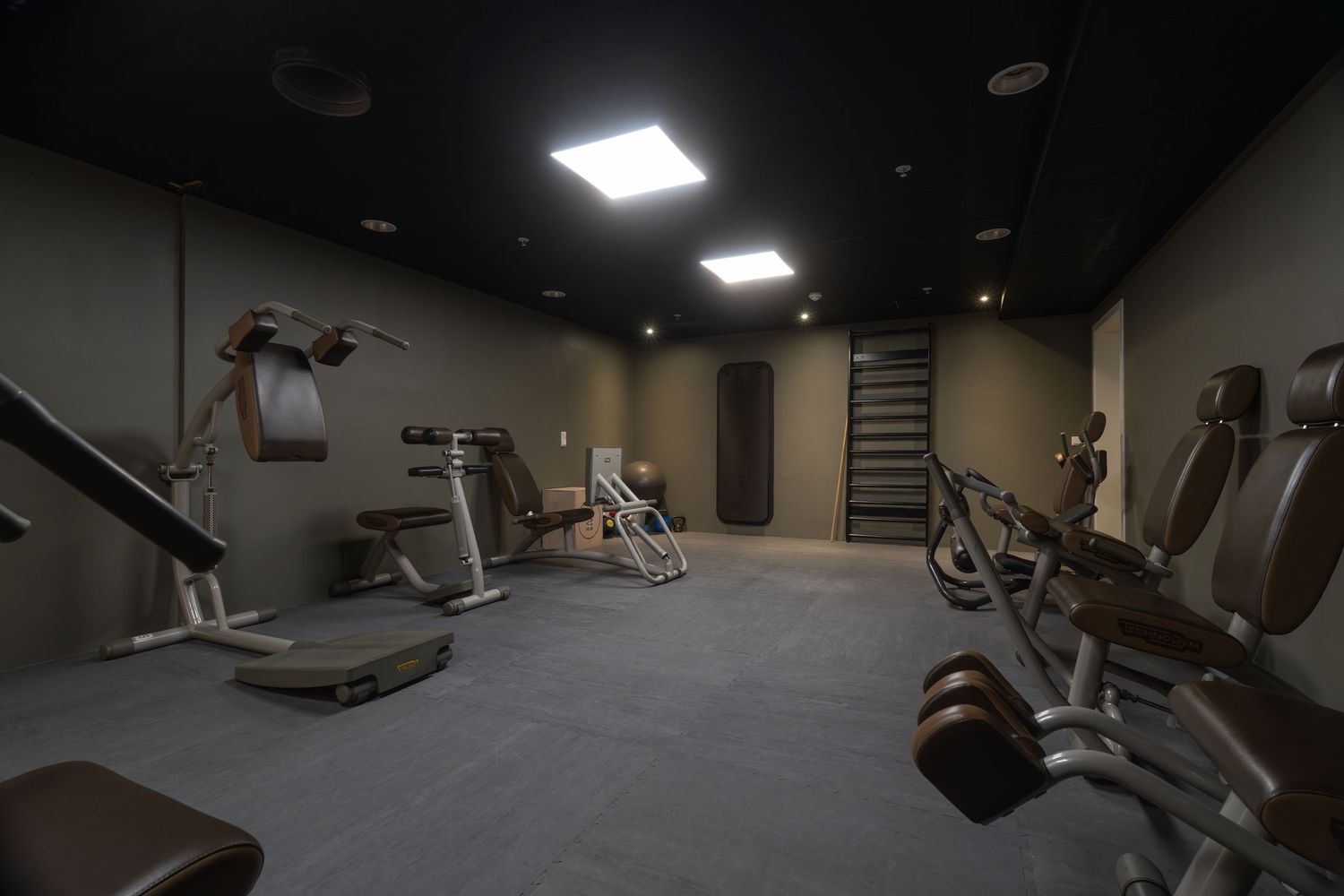 Dark fitness room interior showing silhouettes of various gym equipment and machines.
